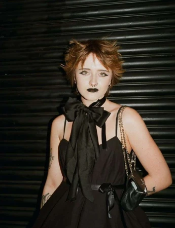 Portrait of a young person with short reddish-brown hair, dark makeup, and a large black bow choker standing against a dark corrugated metal shutter.
