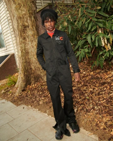 Young Black man in a black jumpsuit and beret posing outdoors near a tree-lined walkway with fallen leaves