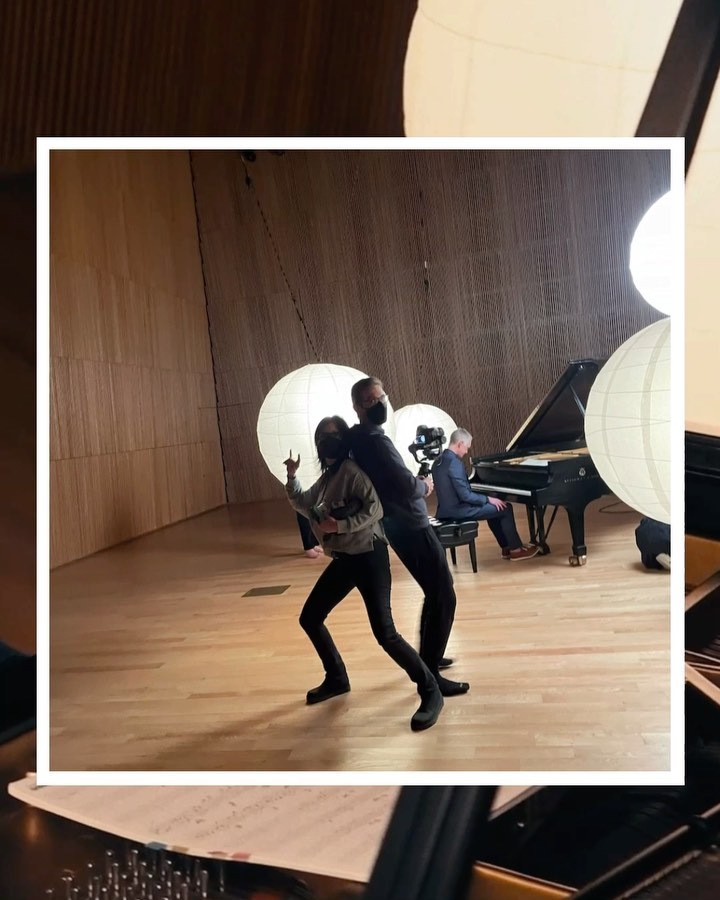 Two people pose in a concert hall, one in a mask holding up two fingers, piano player, stage lighting, and musical performance space.