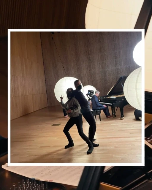 Two people pose in a concert hall, one in a mask holding up two fingers, piano player, stage lighting, and musical performance space.