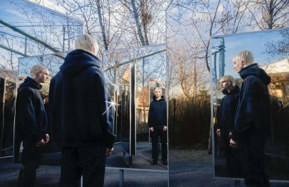 Multiple mirrored images of a young adult with bleached blonde hair wearing a dark hoodie, posing outdoors in an urban setting.