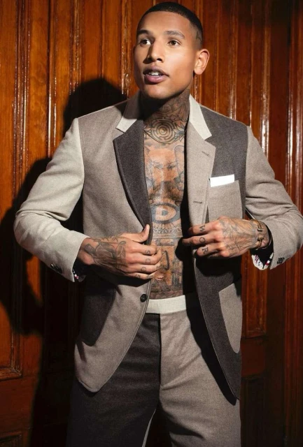 Tattooed adult male posing in an open two-tone suit with a wood panel background. He is looking up and to the left of the frame.