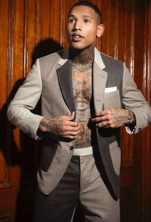 Tattooed adult male posing in an open two-tone suit with a wood panel background. He is looking up and to the left of the frame.