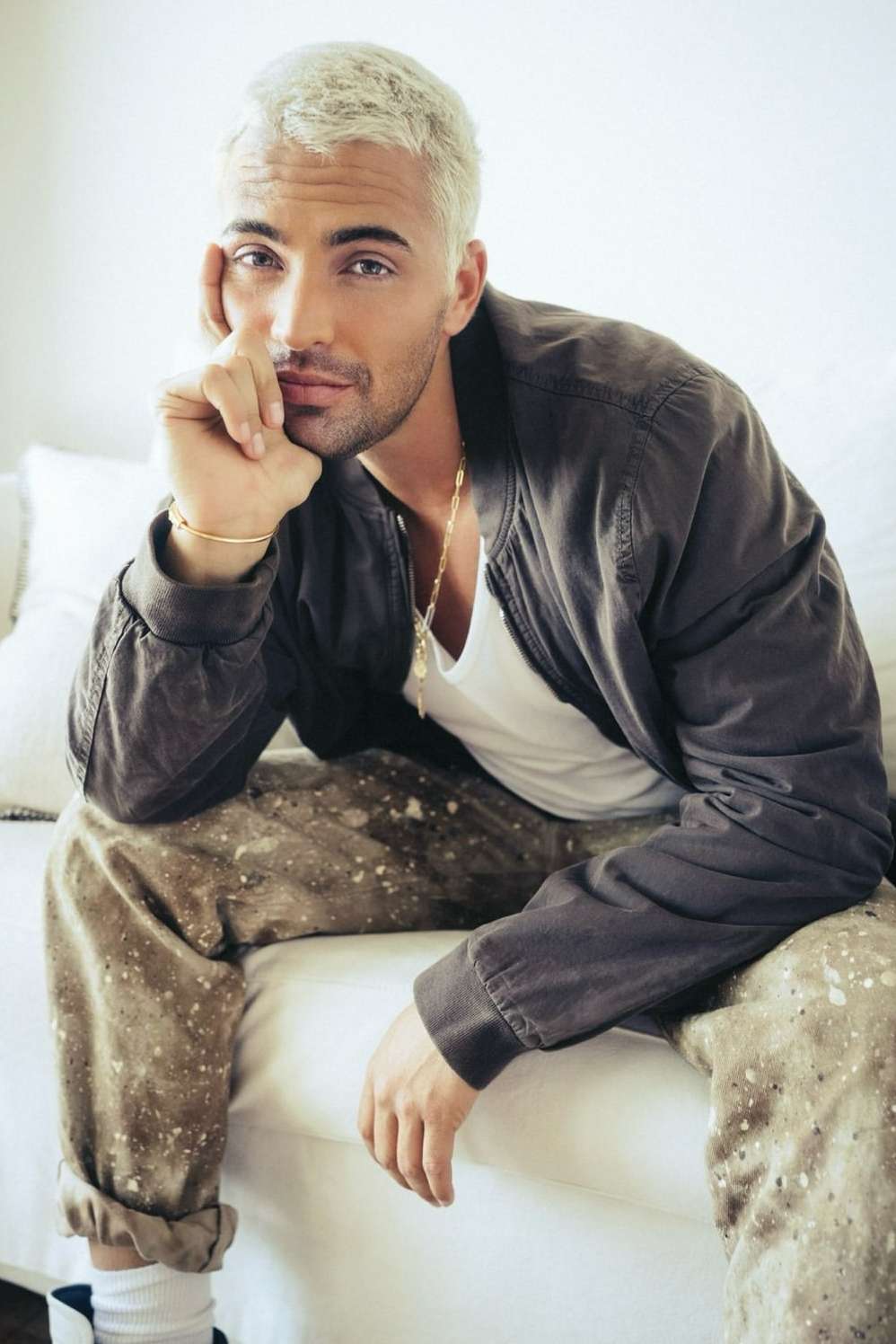 Portrait of a platinum-haired adult male model wearing a bomber jacket, t-shirt, and patterned pants while seated indoors.