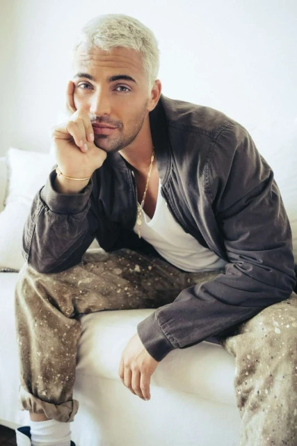 Portrait of a platinum-haired adult male model wearing a bomber jacket, t-shirt, and patterned pants while seated indoors.