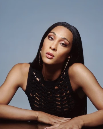 Portrait of a woman with long dark hair wearing a black knit top and hoop earrings against a grey background.