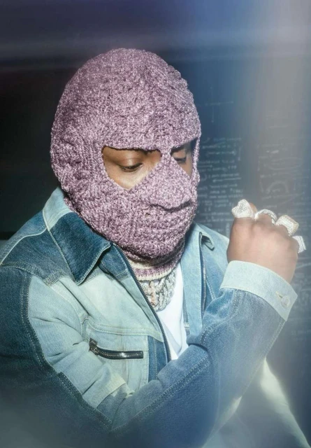Portrait of a person wearing a knit balaclava and denim jacket, accessorized with a chunky chain and multiple rings, against a blurred background.