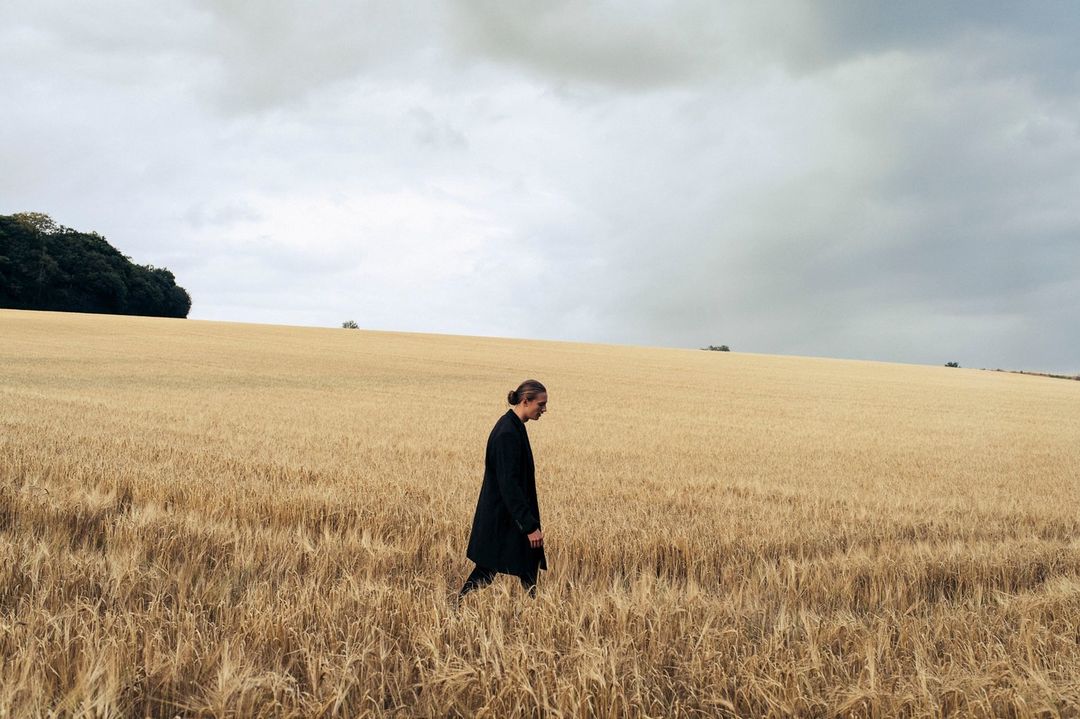 An adult in a black coat walks through a field of golden wheat under an overcast sky, creating a solitary and contemplative mood.