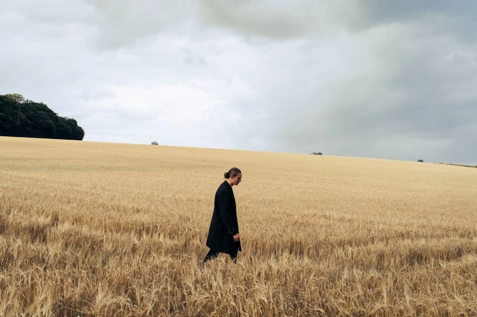 An adult in a black coat walks through a field of golden wheat under an overcast sky, creating a solitary and contemplative mood.