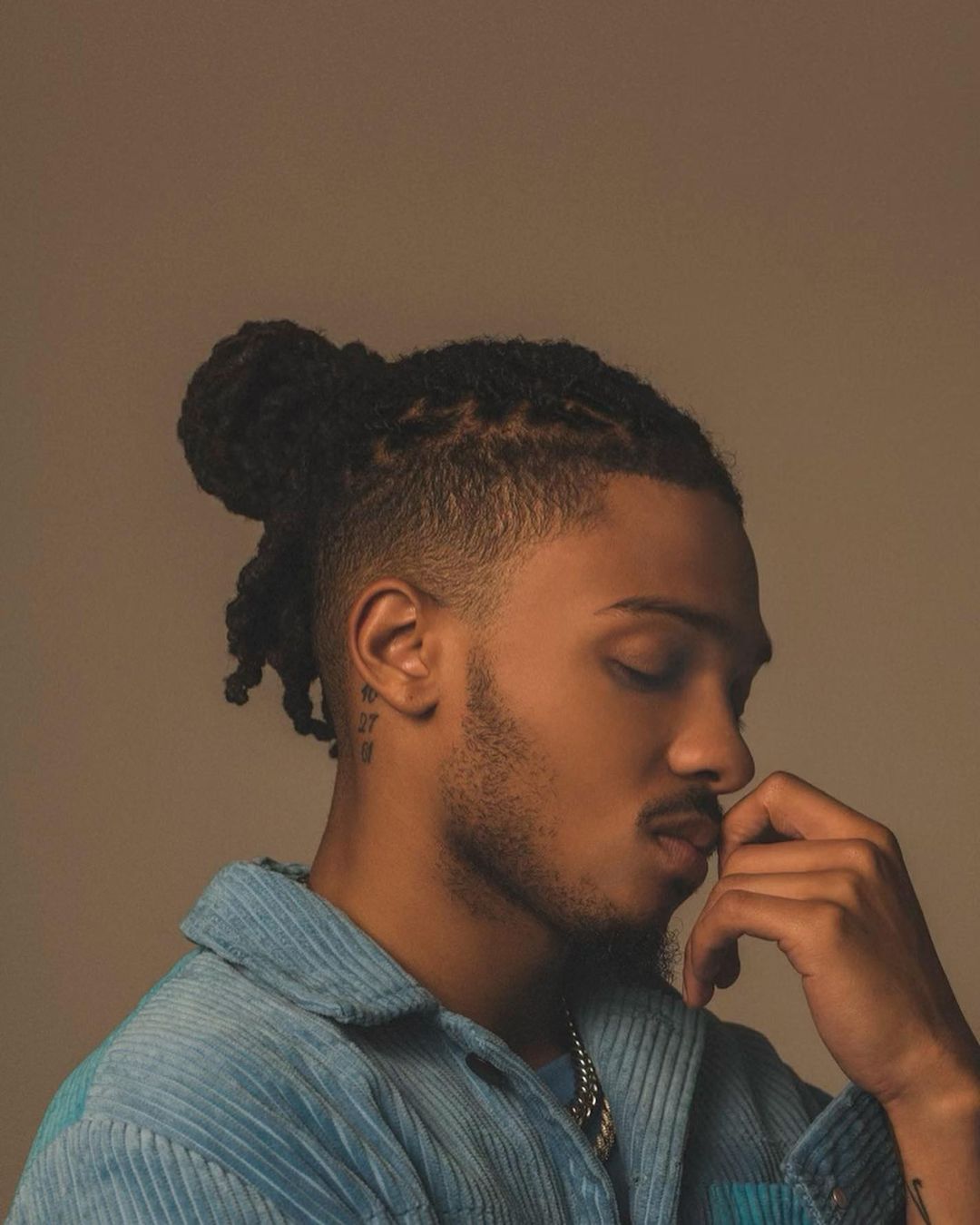 Close-up portrait of a Black male with dreadlocks in a high bun, beard, and a light blue corduroy shirt against a neutral background.