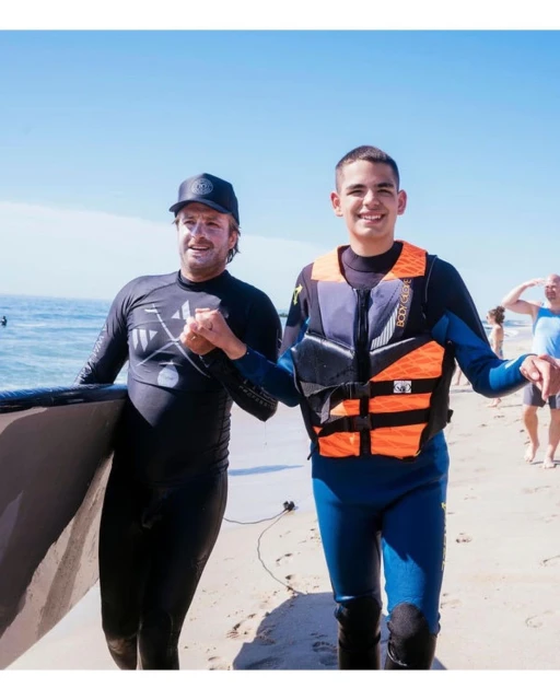 Two males on a beach, one wearing a life jacket and the other carrying a surfboard, enjoying a sunny day near the ocean.