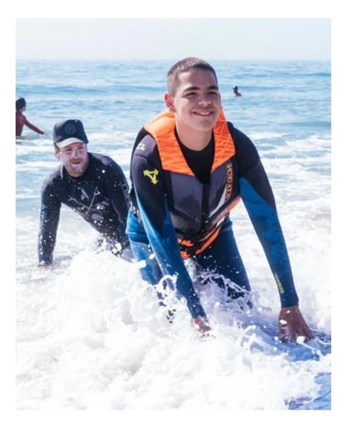Two people in wetsuits on a beach. A young man wearing a life vest kneels in the surf with another man standing behind him.