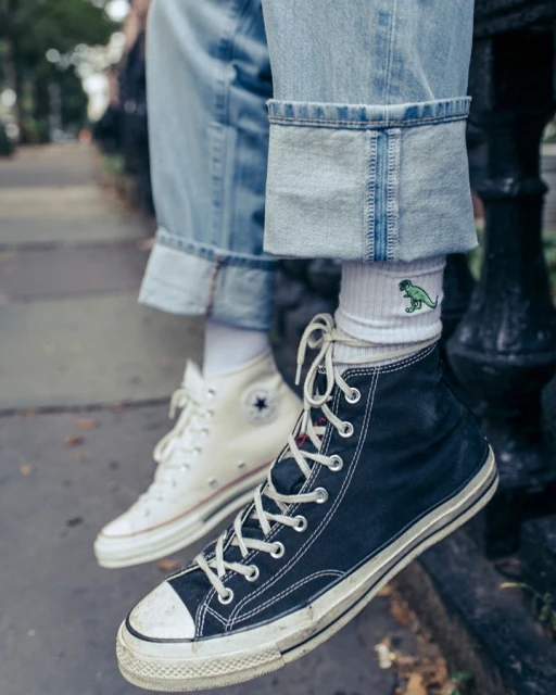 High-top sneakers in black and white colors are worn with jeans and dinosaur socks on a concrete surface outdoors.