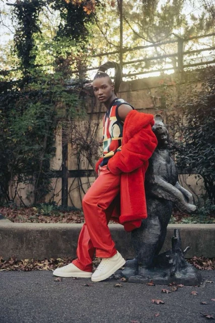 Fashionable non-binary person in a colorful top and red pants and jacket leaning against a sculpture in an outdoor setting.