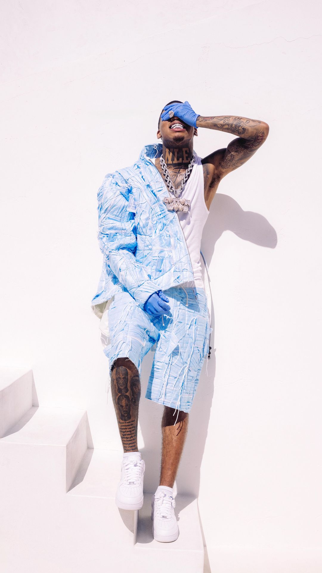 Man in light blue patterned shorts and jacket covers his face, posing against a white background with geometric shapes and shadows.