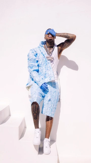 Man in light blue patterned shorts and jacket covers his face, posing against a white background with geometric shapes and shadows.