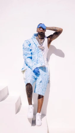 Man in light blue patterned shorts and jacket covers his face, posing against a white background with geometric shapes and shadows.