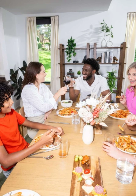 Diverse group of friends and family gather around a wooden table for a shared meal with pasta, appetizers, and wine in a bright room.
