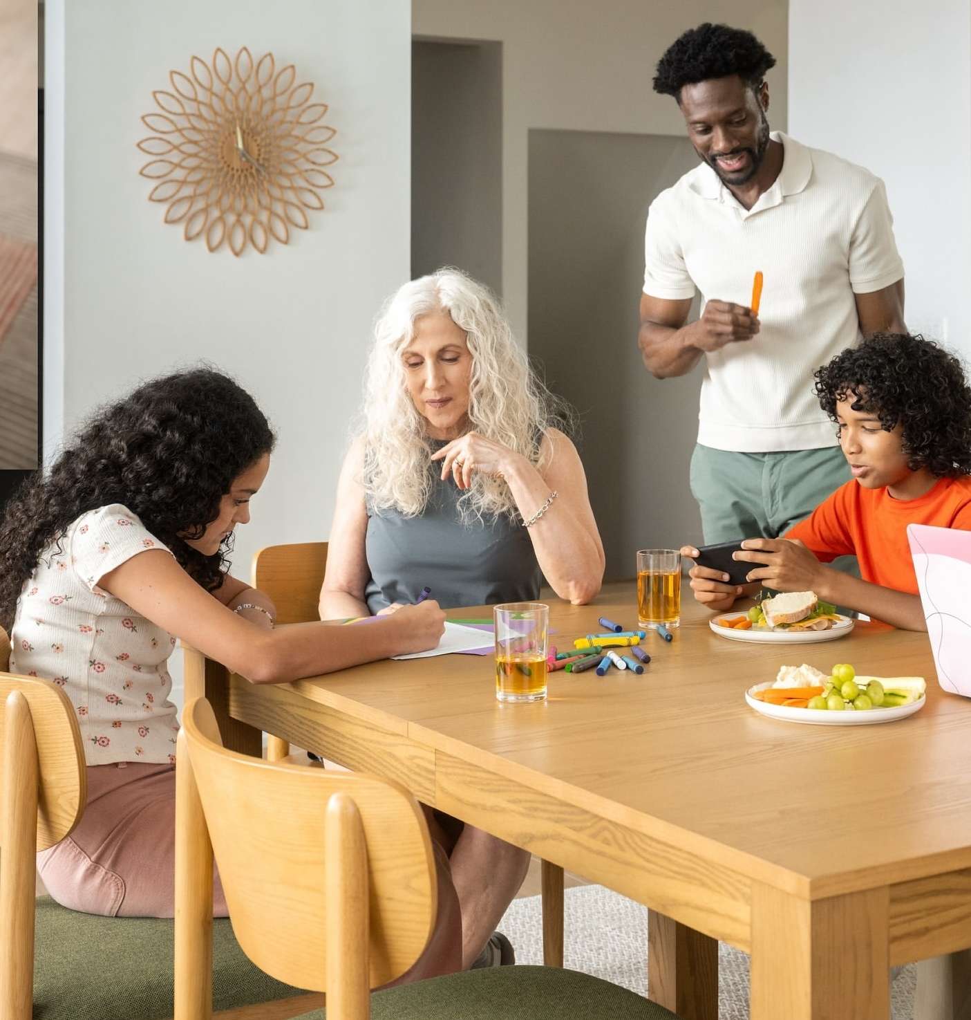 A family of four gathers around a wooden table in a brightly lit interior. Two children are engaged with activities while two adults are present.