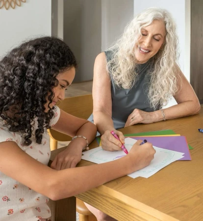 Smiling senior woman helps girl with art project on wooden table with colored paper and markers indoors in daylight, creating art together.