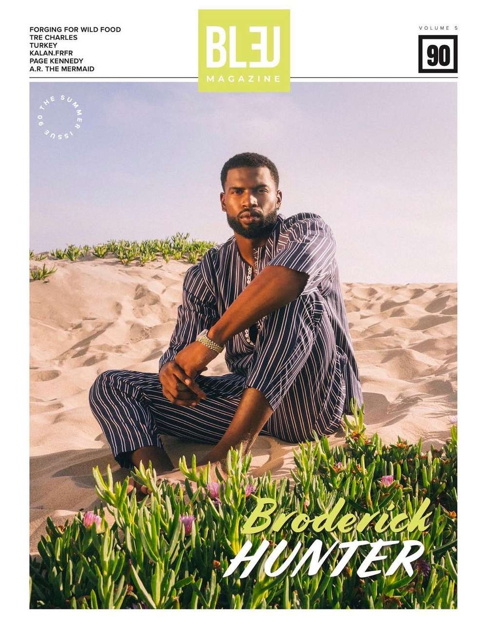 Black man with a beard seated in sand dunes wearing a striped two-piece outfit, photographed by Andrew Zaeh.