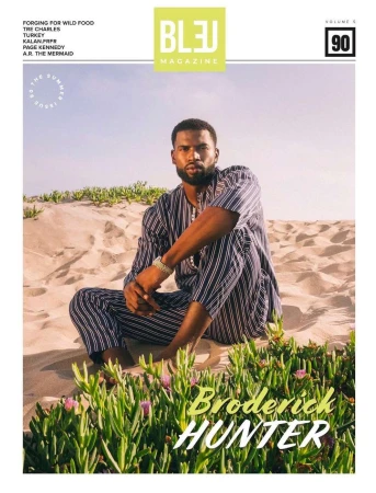 Black man with a beard seated in sand dunes wearing a striped two-piece outfit, photographed by Andrew Zaeh.