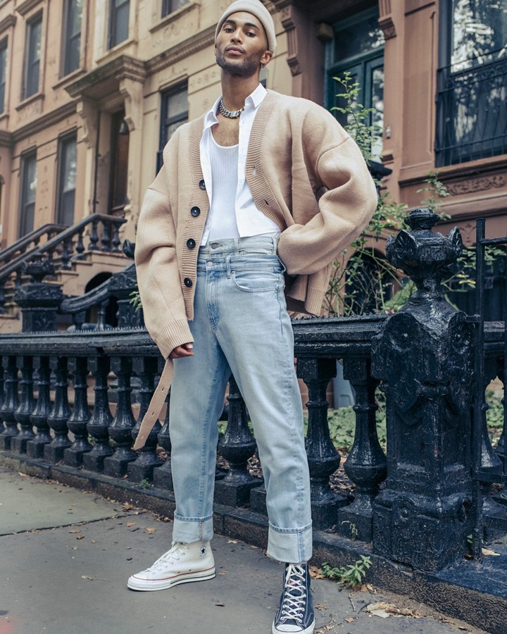 Man in beige cardigan, white tank top, and light wash jeans stands in front of a brownstone building with wrought iron details.