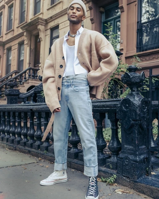 Man in beige cardigan, white tank top, and light wash jeans stands in front of a brownstone building with wrought iron details.