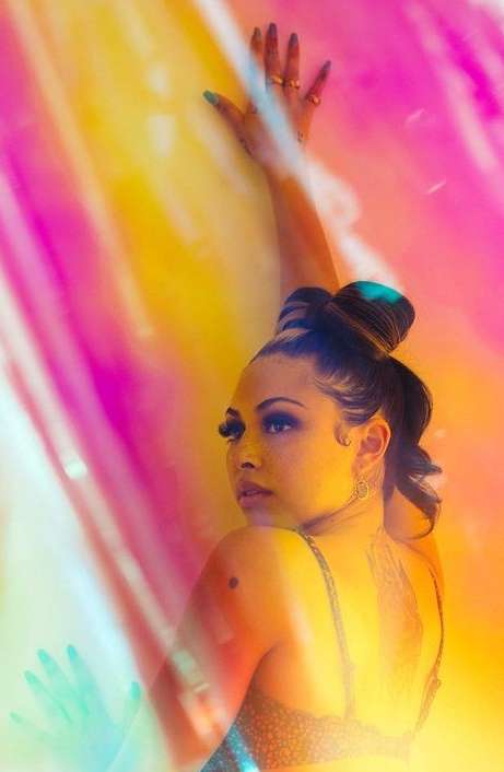 Adult female poses in a colorful, light-filled setting, showcasing an updo hairstyle, jewelry, and a bra top against a vibrant background.