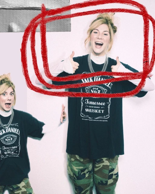 A young blonde woman smiles, wears a Jack Daniel's t-shirt and camo pants, and gives two thumbs up in a bright, indoor setting.