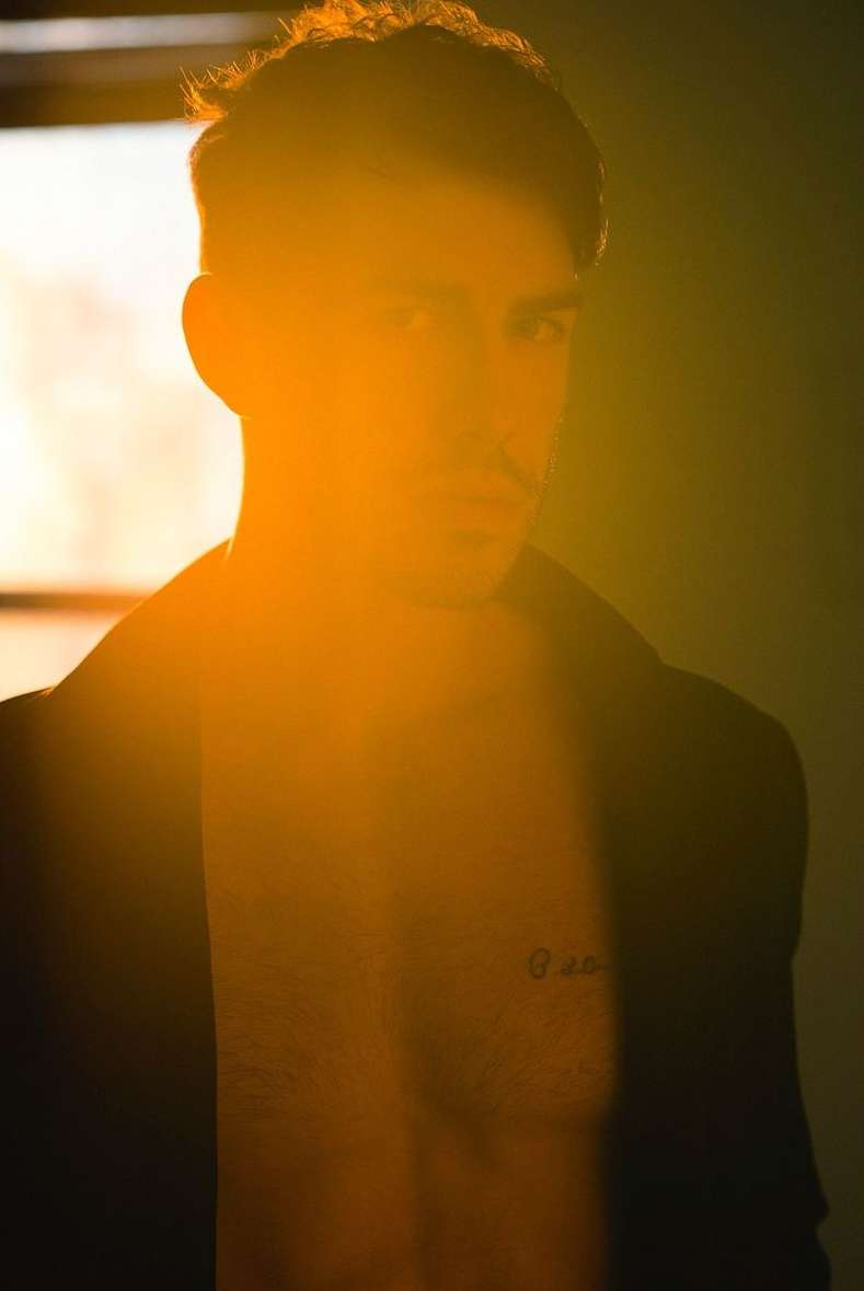 Portrait of a shirtless adult male with a jacket and dark hair, strongly backlit with golden light, revealing chest tattoo.