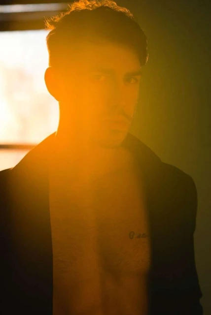 Portrait of a shirtless adult male with a jacket and dark hair, strongly backlit with golden light, revealing chest tattoo.
