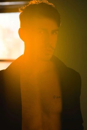 Portrait of a shirtless adult male with a jacket and dark hair, strongly backlit with golden light, revealing chest tattoo.
