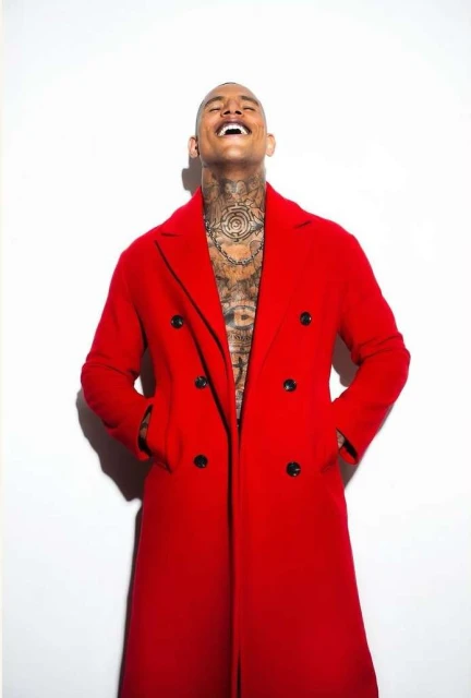 Smiling bald adult male in an open bright red overcoat showing chest and neck tattoos against a white backdrop in a studio shot.