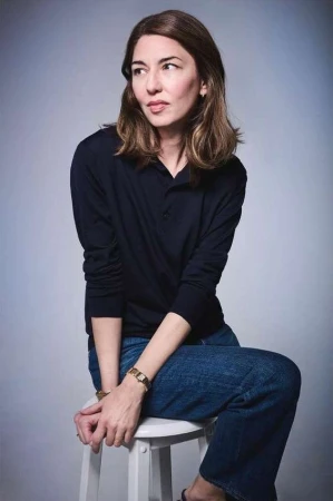 Portrait of a woman seated on a white stool wearing a dark polo shirt and denim jeans against a grey background.