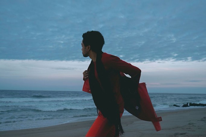 A man in a red outfit and dark scarf walking on a sandy beach near ocean waves under a cloudy sky.