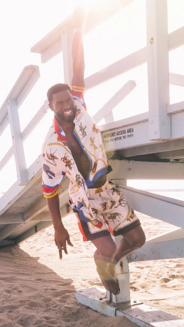 Athletic Black man in patterned shirt and shorts hangs from lifeguard tower on sunlit beach, smiling with open mouth, barefoot.