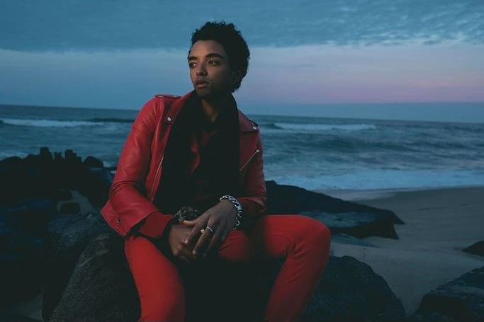 Man in red leather jacket and pants sits on rocks by the ocean at dusk, gazing out at the sea with a contemplative expression.