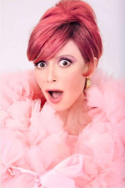 Adult woman with bright pink hair and heavy makeup, wearing a frilly pink top with a surprised facial expression in a studio.