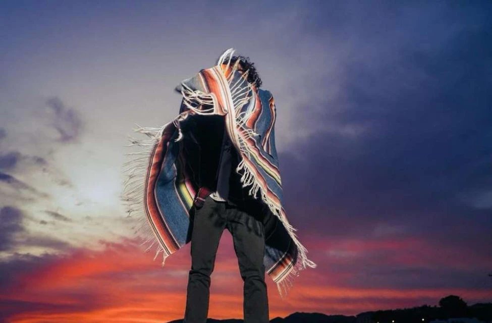 Person wrapped in fringed blanket standing against a gradient sky with red and blue hues in an outdoor landscape. Full shot.