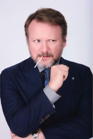 A man in a navy blue suit biting a small white stick against a plain white background, photographed by Andrew Zaeh.