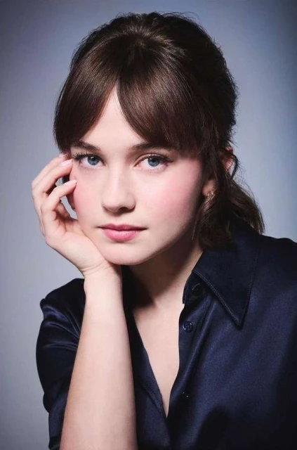 Close-up portrait of a young woman with brown hair, wearing a navy blue blouse, resting her face on her hand in a studio setting.