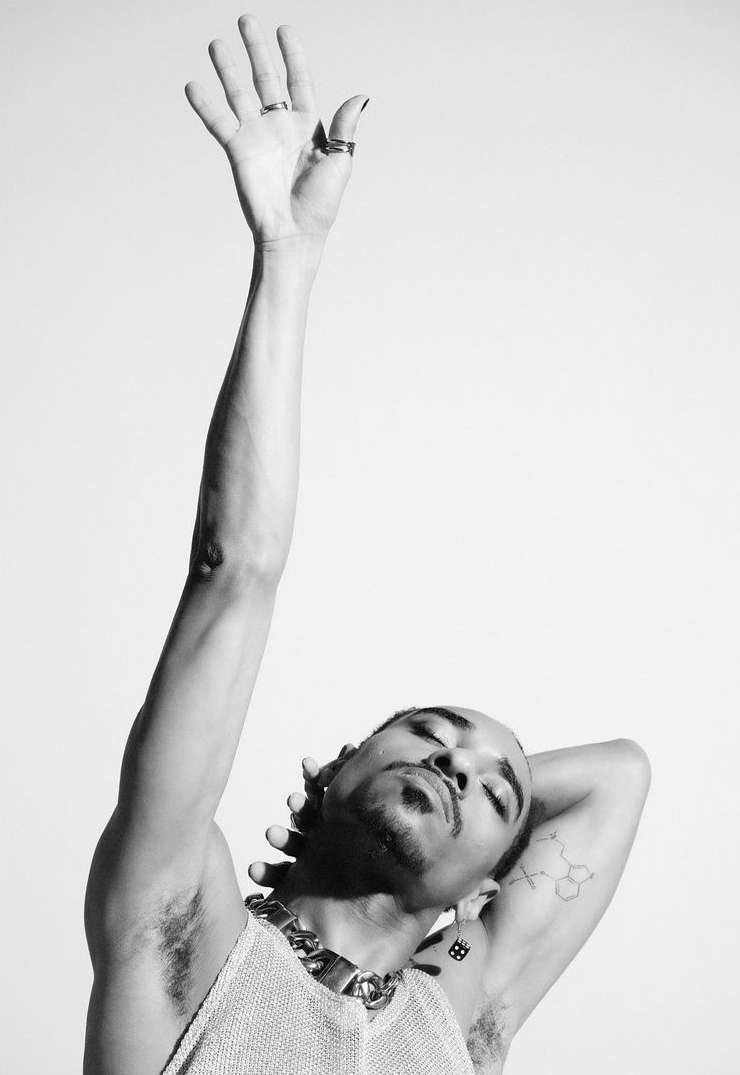 Monochrome portrait of a man with arm raised, eyes closed, wearing a chain necklace against a plain white background.