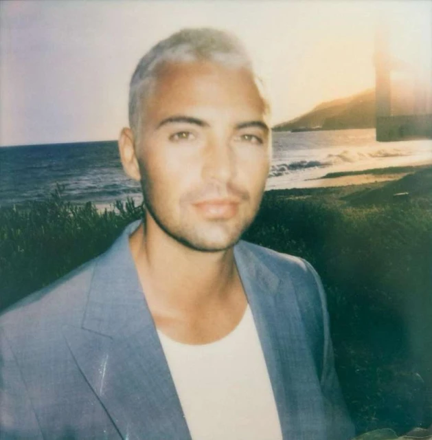 Portrait of an adult male with short silver hair wearing a blue suit jacket, posing on a beach with ocean and green plants in the background.