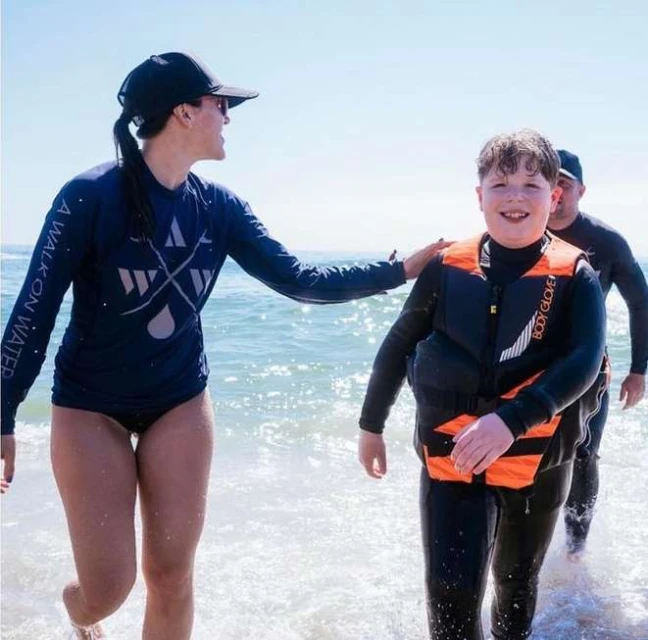 Smiling child in life vest walks on beach with two adults and ocean waves, medium shot, sunny day. Life jacket, swimwear, water sport.