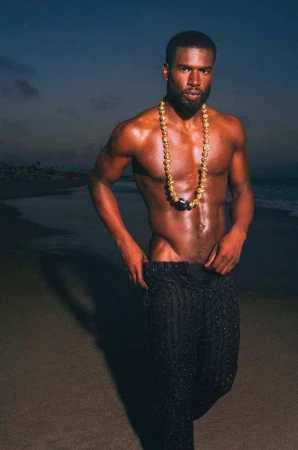 Shirtless man wearing beaded necklace and dark textured pants walking on a beach at dusk