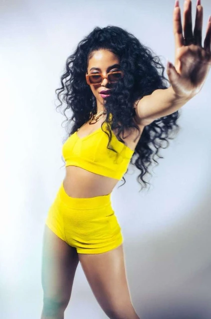 A young mixed-race woman with dark curly hair and sunglasses poses in yellow activewear against a gradient white backdrop indoors.