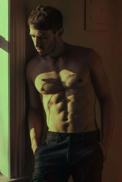 Shirtless man poses by window. Male fitness model with six-pack abs in black pants leans against window in dramatic light. Studio shot.
