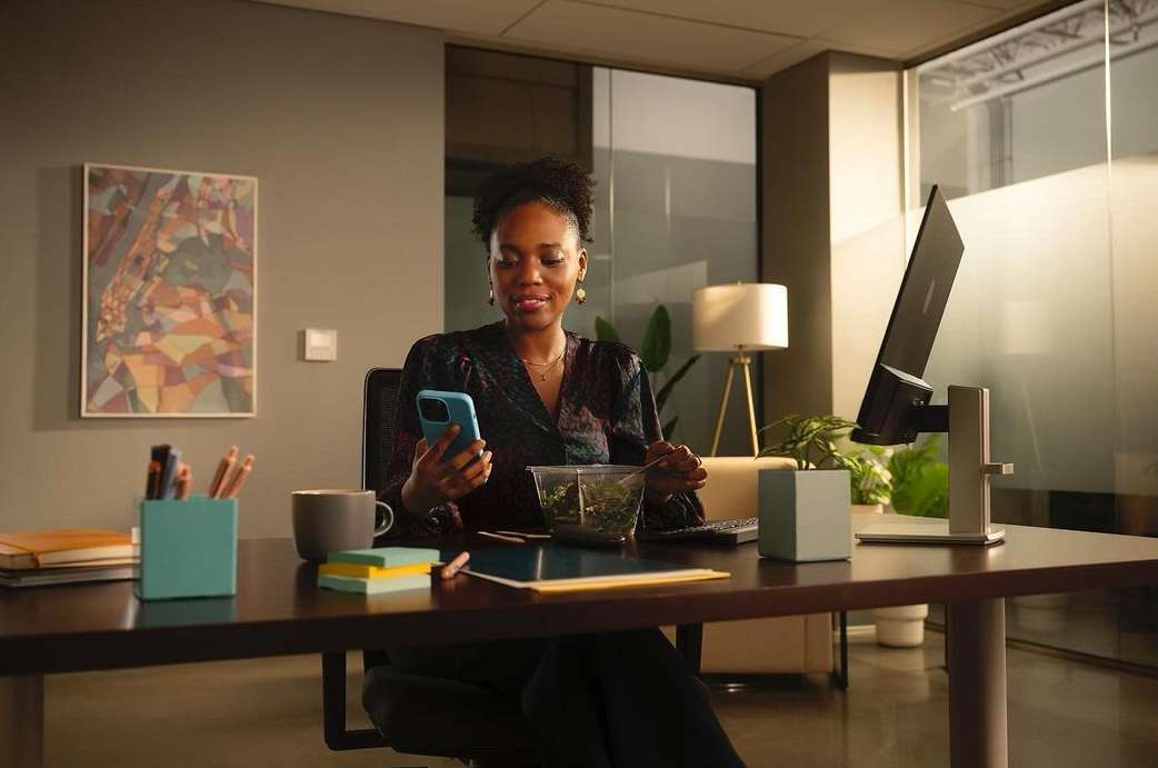 Businesswoman checks her phone while eating salad at office desk. Modern workspace includes computer, lamp, decor, and office supplies.