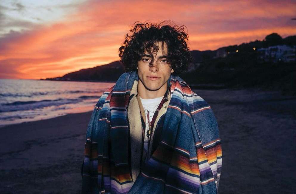 A young man with curly dark hair wrapped in a colorful striped blanket on a sandy beach at sunset.